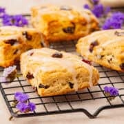 Four cranberry glazed scones with dried fruit rest on a cooling rack, surrounded by small purple flowers.