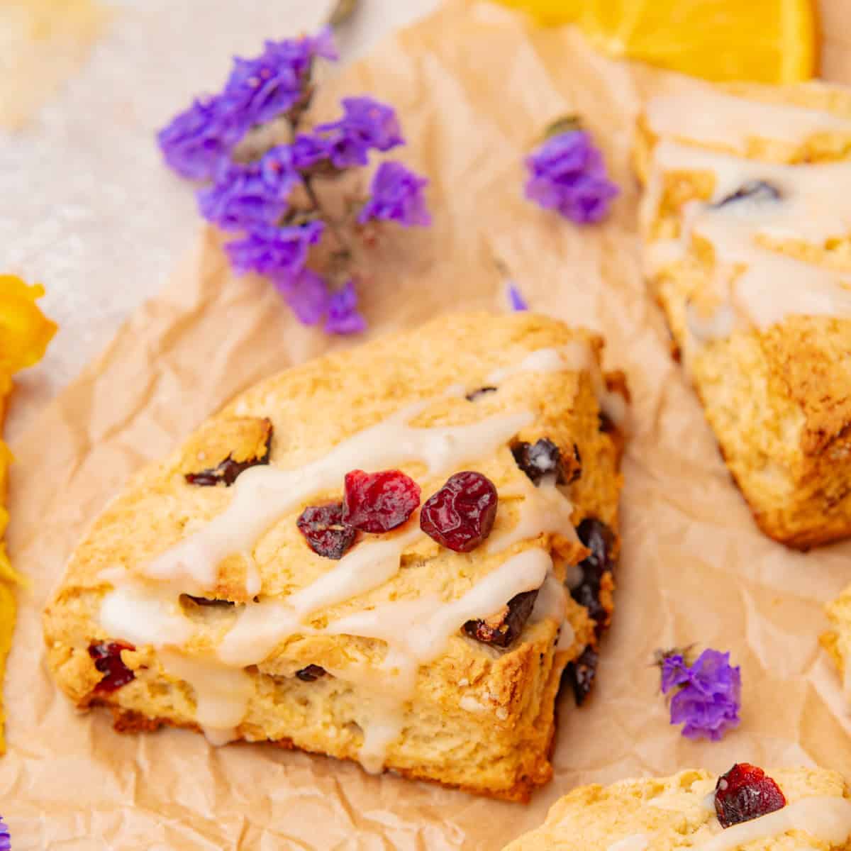 A triangular cranberry glazed scone with white icing drizzle sits on brown parchment paper, surrounded by small purple flowers.