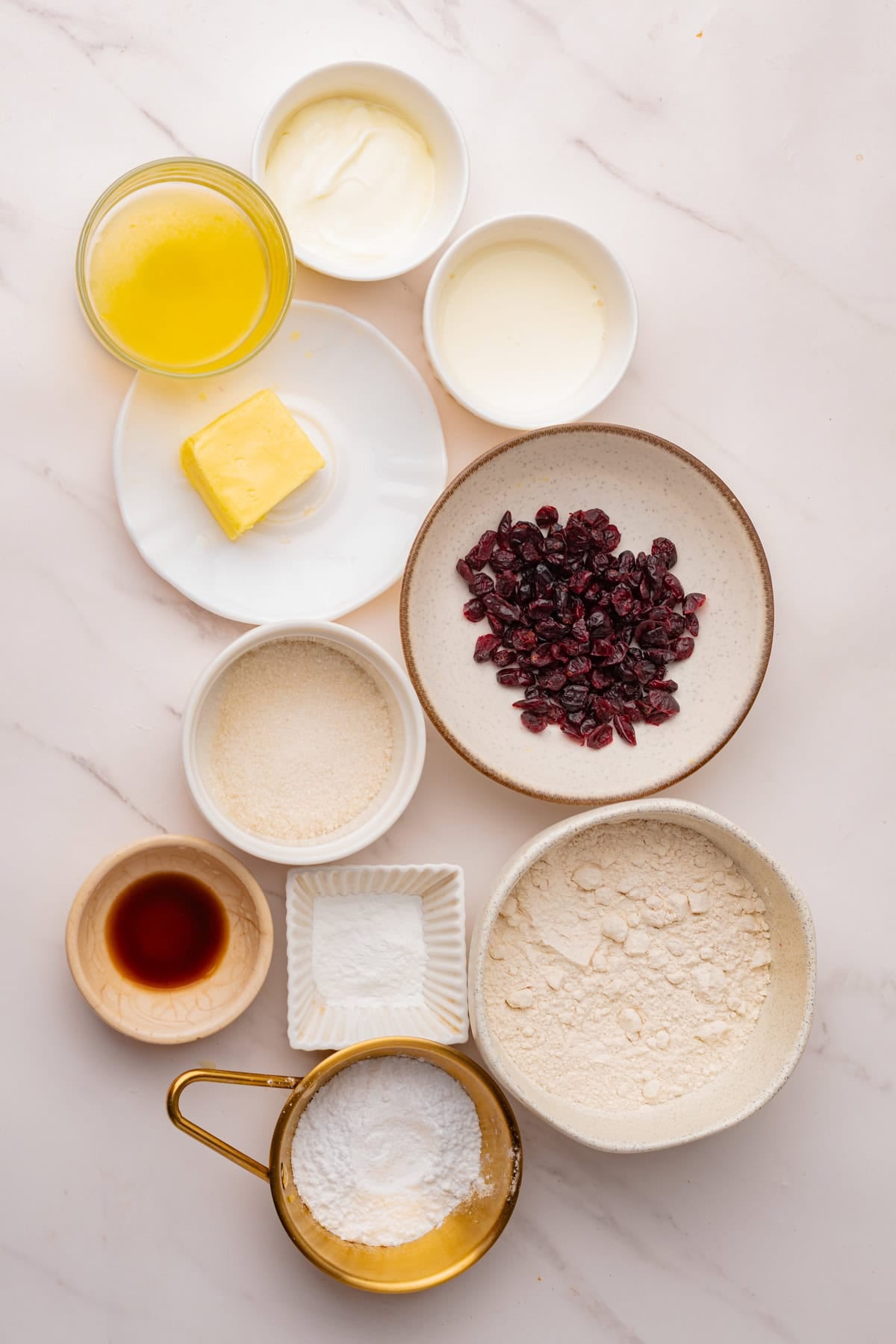 Various baking ingredients arranged on a marble surface, including flour, sugar, powdered sugar, butter, vanilla, yogurt, and dried cranberries—everything you need to make delicious cranberry glazed scones.