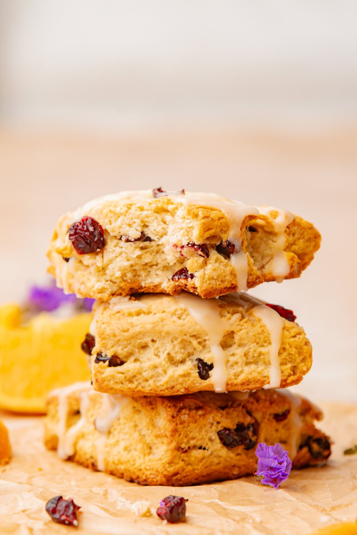 Three cranberry glazed scones are stacked on parchment paper, garnished with small purple flowers and cranberries.