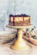 6inch blackberry cheesecake on yellow cake plate with plates in background.