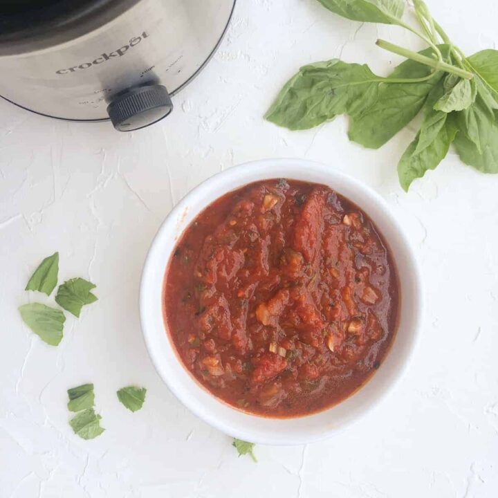 bowl of homemade slow cooker marina sauce and basil leaves with crockpot in background.