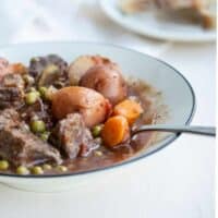 bowl of beef stew with peas, carrots, and potatoes with bread in background.