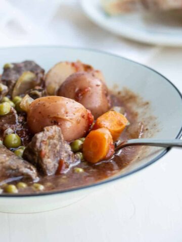 bowl of beef stew with peas, carrots, and potatoes with bread in background.
