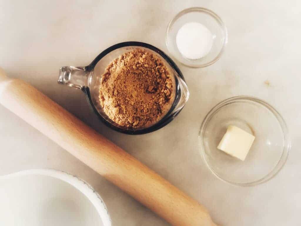 Butter, sugar and crushed graham crackers on table next to a rolling pin.
