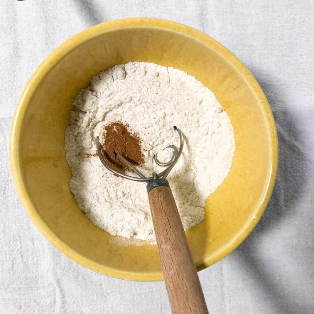 pumpkin muffins dry ingredients in yellow bowl and dough whisk on white cloth.
