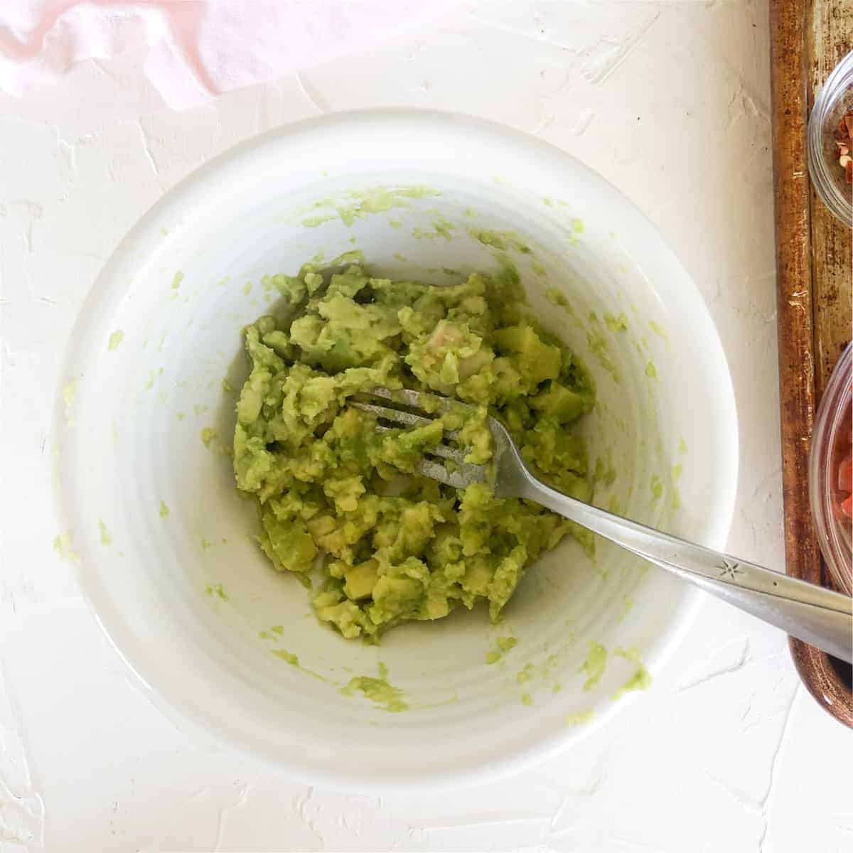 avocado cubes mashed with fork in a white bowl.