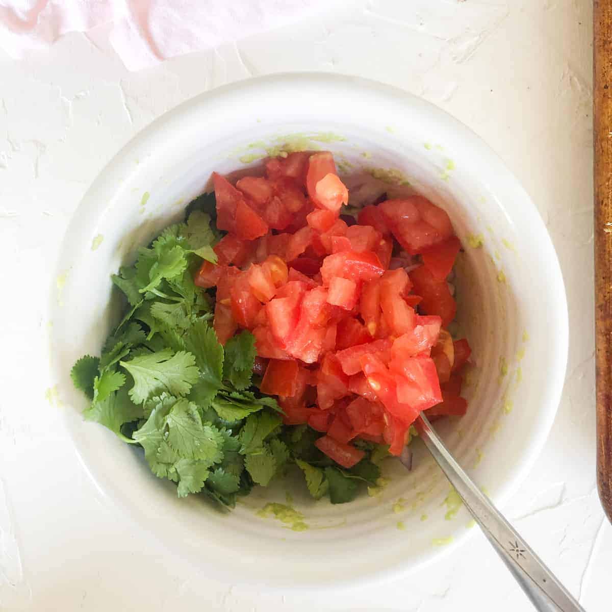 chopped cilantro leaves and diced tomatoes on top of mashed avocado in white bowl.