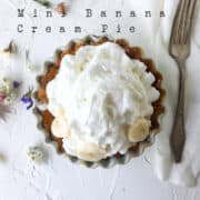 whipped cream on top of a small banana cream pie with flowers scattered on table.