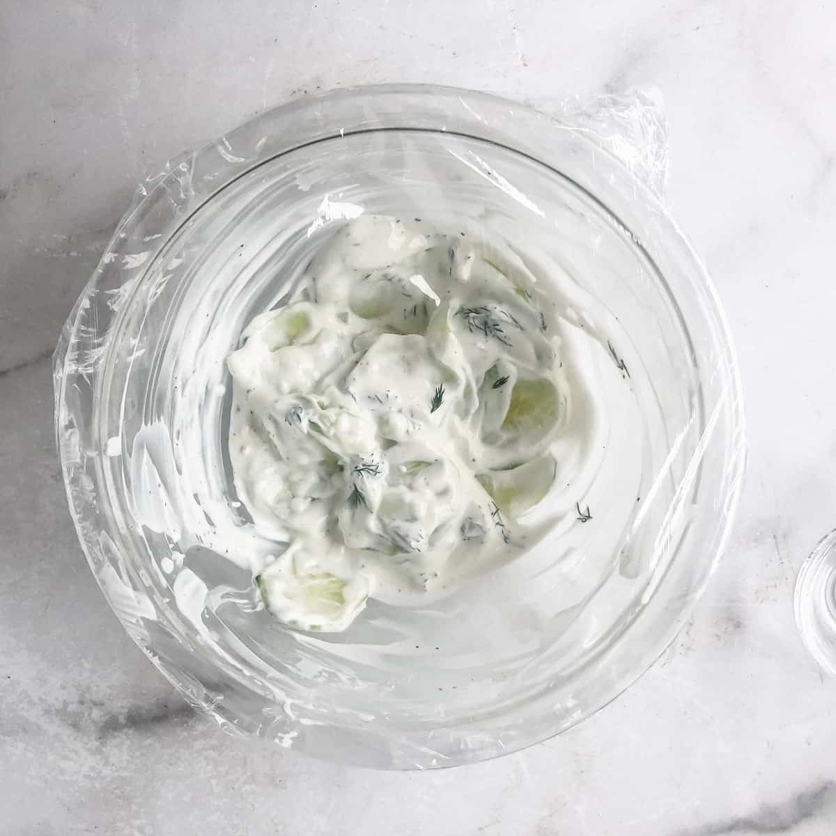 Covered bowl of cucumber and sour cream with dill.