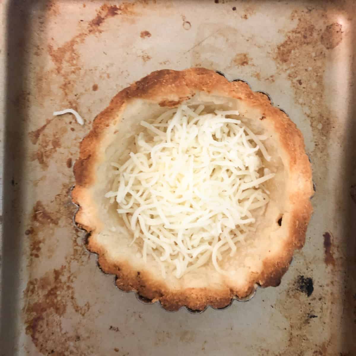 Shredded cheese nestled in a small baked pie crust sits on a stained baking sheet.