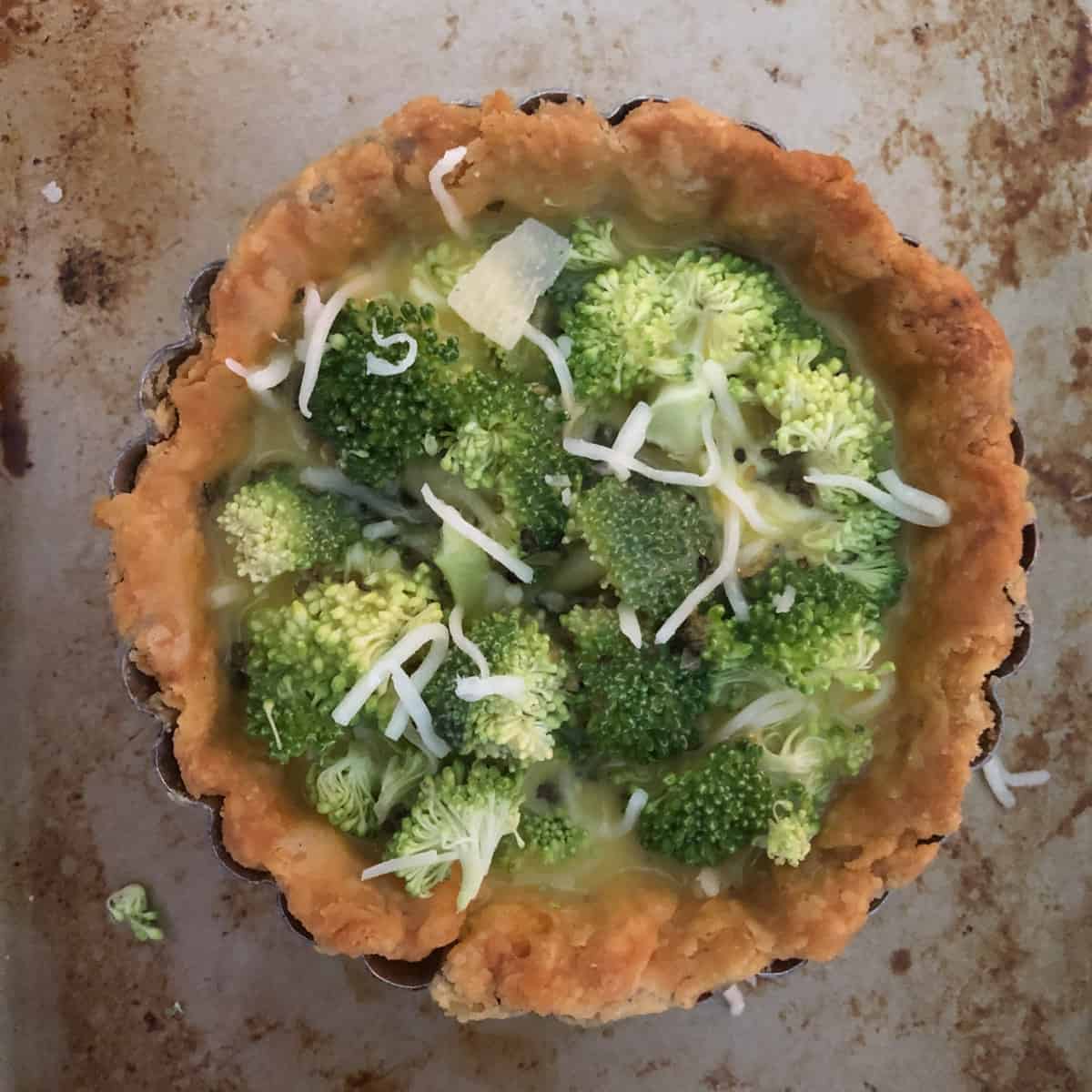 Unbaked mini broccoli quiche for one with cheese in a golden crust on a baking tray.