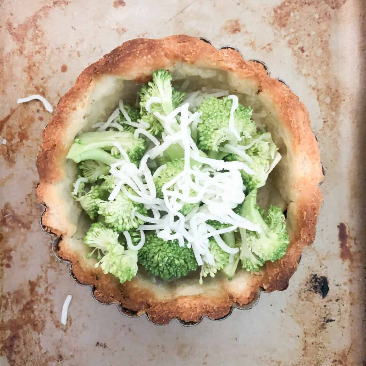 Blind baked quiche crust filled with raw broccoli florets and shredded cheese on a baking sheet.