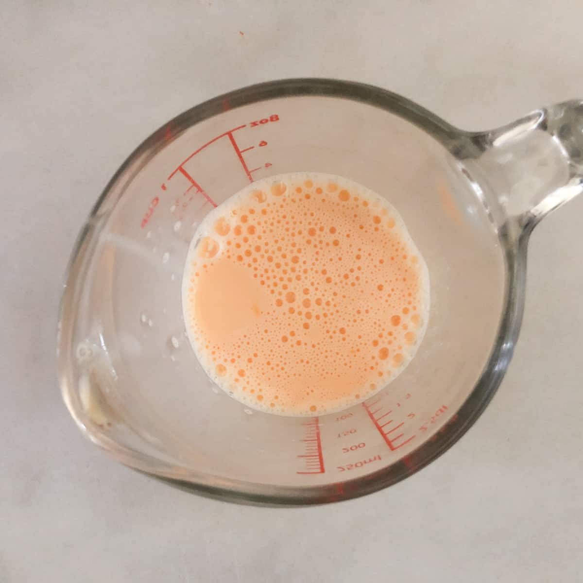 A glass measuring cup with a small amount milk and egg for preparing quiche for one, sits on a light surface.