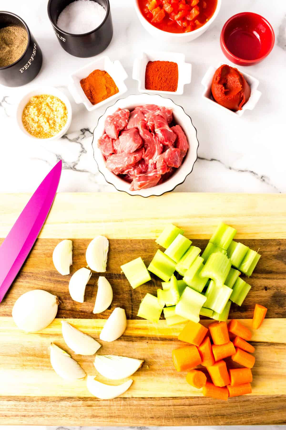 Chopped onion, celery, and carrot on a cutting board with a knife, plus bowls of raw beef ready for paprika beef stew, spices, tomato paste, garlic, and salt and pepper on a marble countertop.