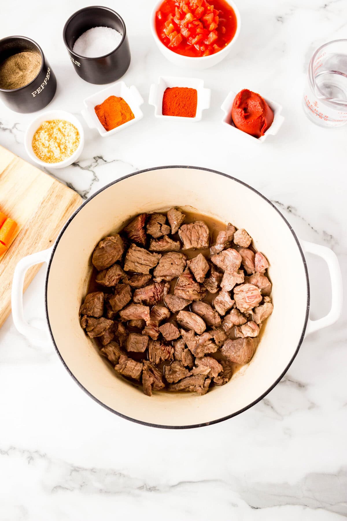 Overhead view of paprika beef stew with browned beef chunks in a white pot, surrounded by small bowls of spices, chopped tomatoes, tomato paste, minced garlic, and seasonings on a marble countertop.