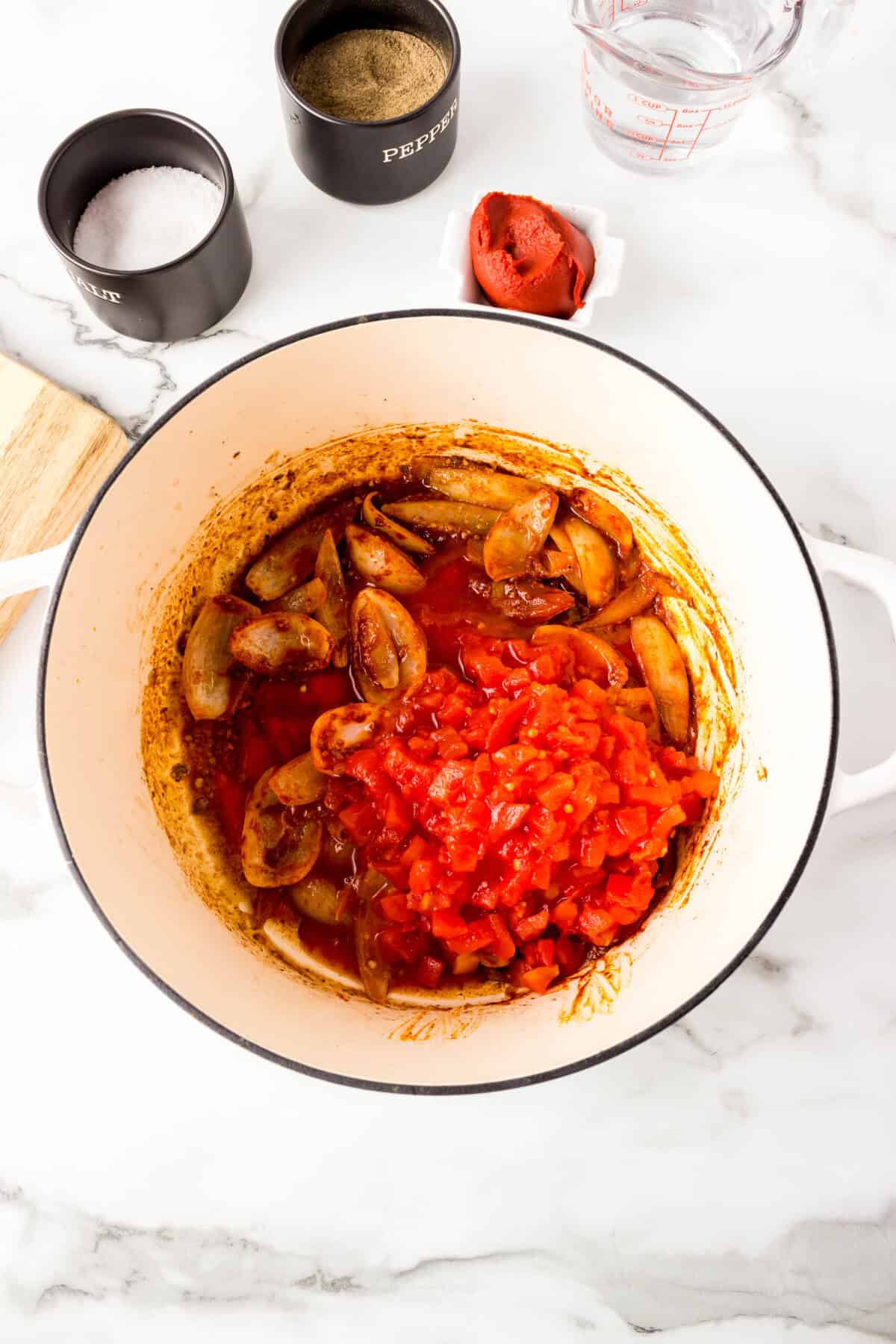 A white pot contains sliced mushrooms, onions, and diced tomatoes in sauce—a savory base for paprika beef stew. Surrounding the pot are containers of salt, pepper, tomato paste, and a measuring cup of water.