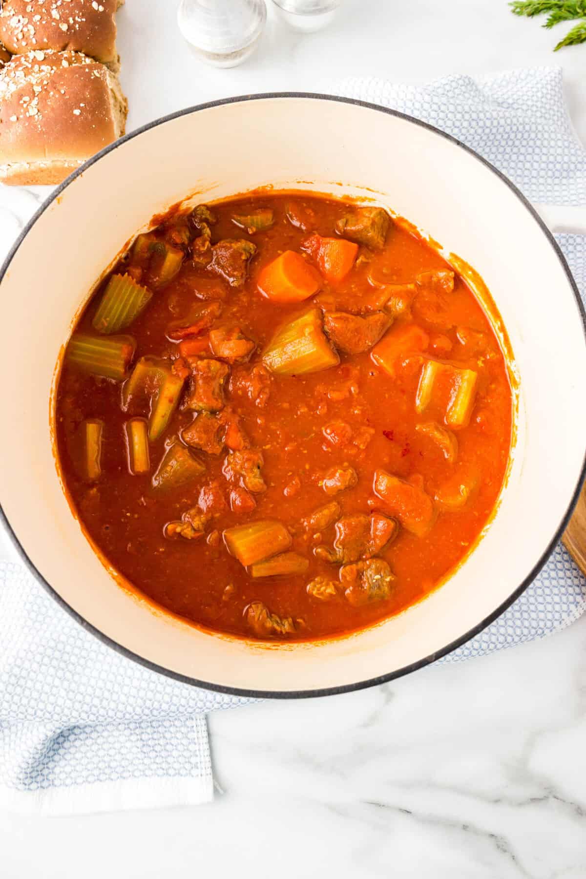 A white pot filled with paprika beef stew featuring chunks of beef, carrots, and celery in a red tomato-based broth, placed on a white surface near sandwich buns.