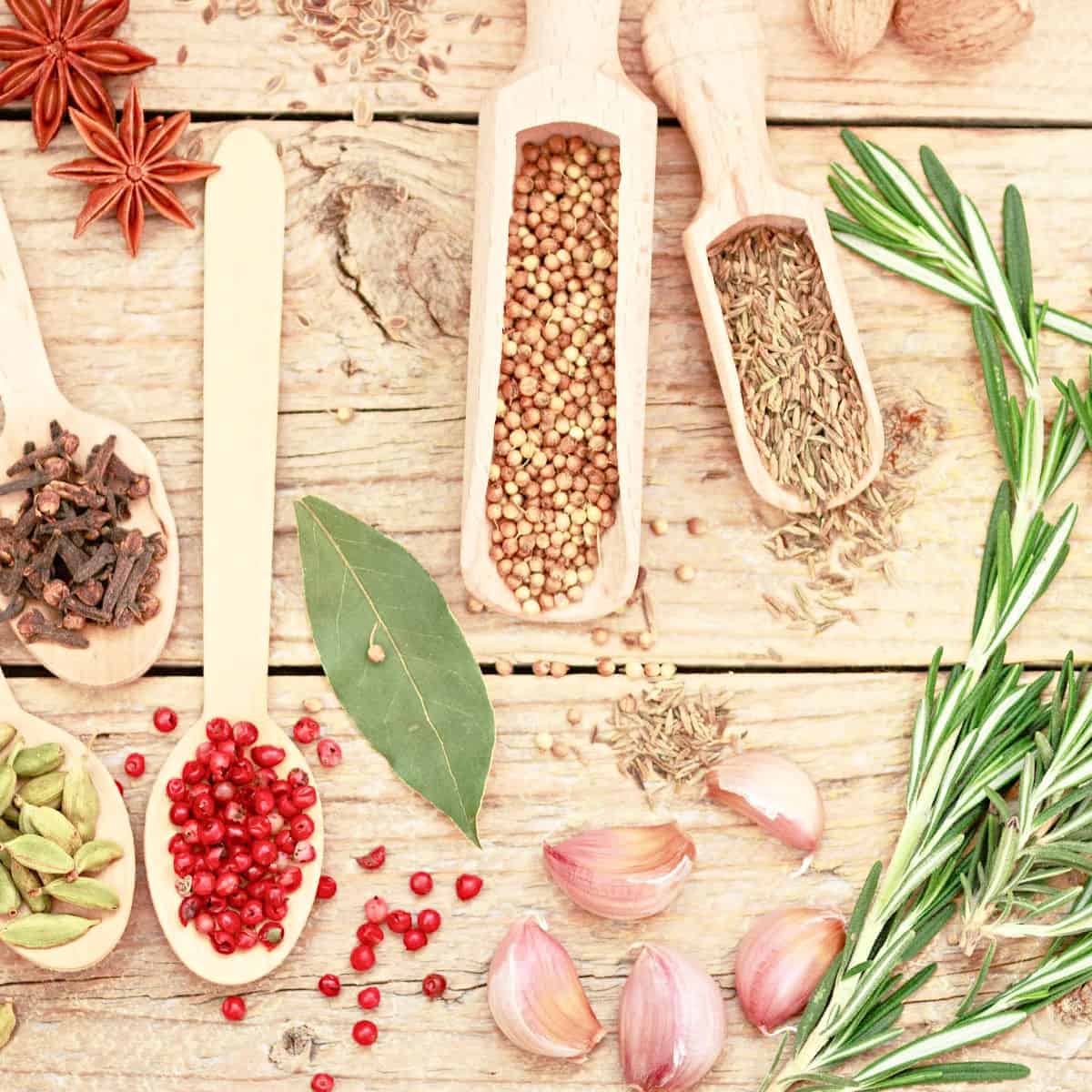 Assorted dried spices in wooden scoops on light wood table.