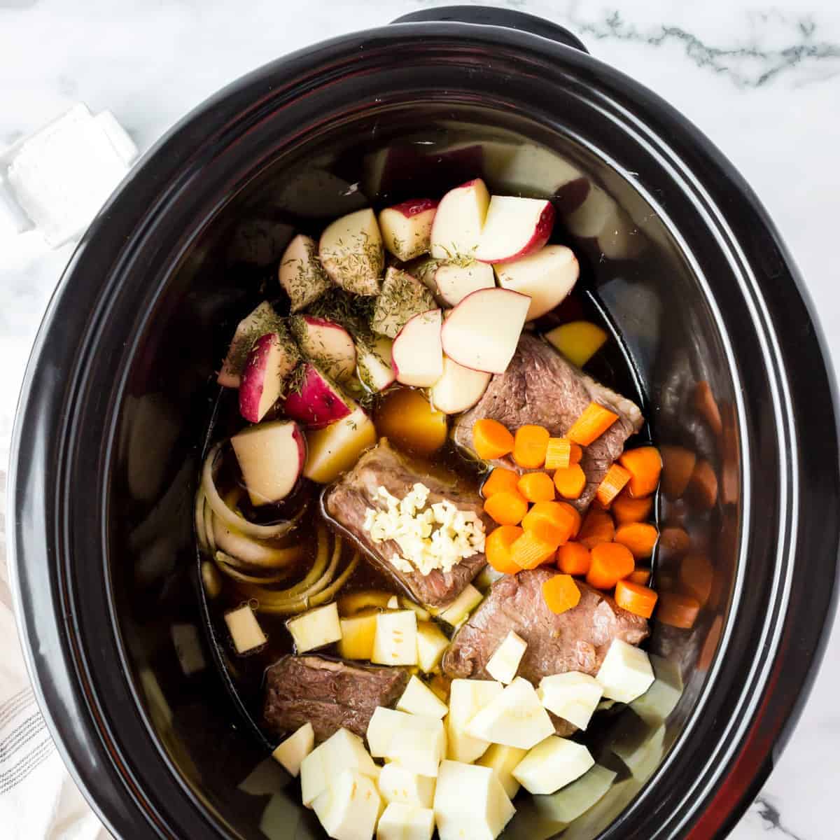 Onions, carrots, potatoes and short ribs in an oval slow cooker.