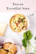 Tortellini stew in a white bowl with fresh spinach on table.
