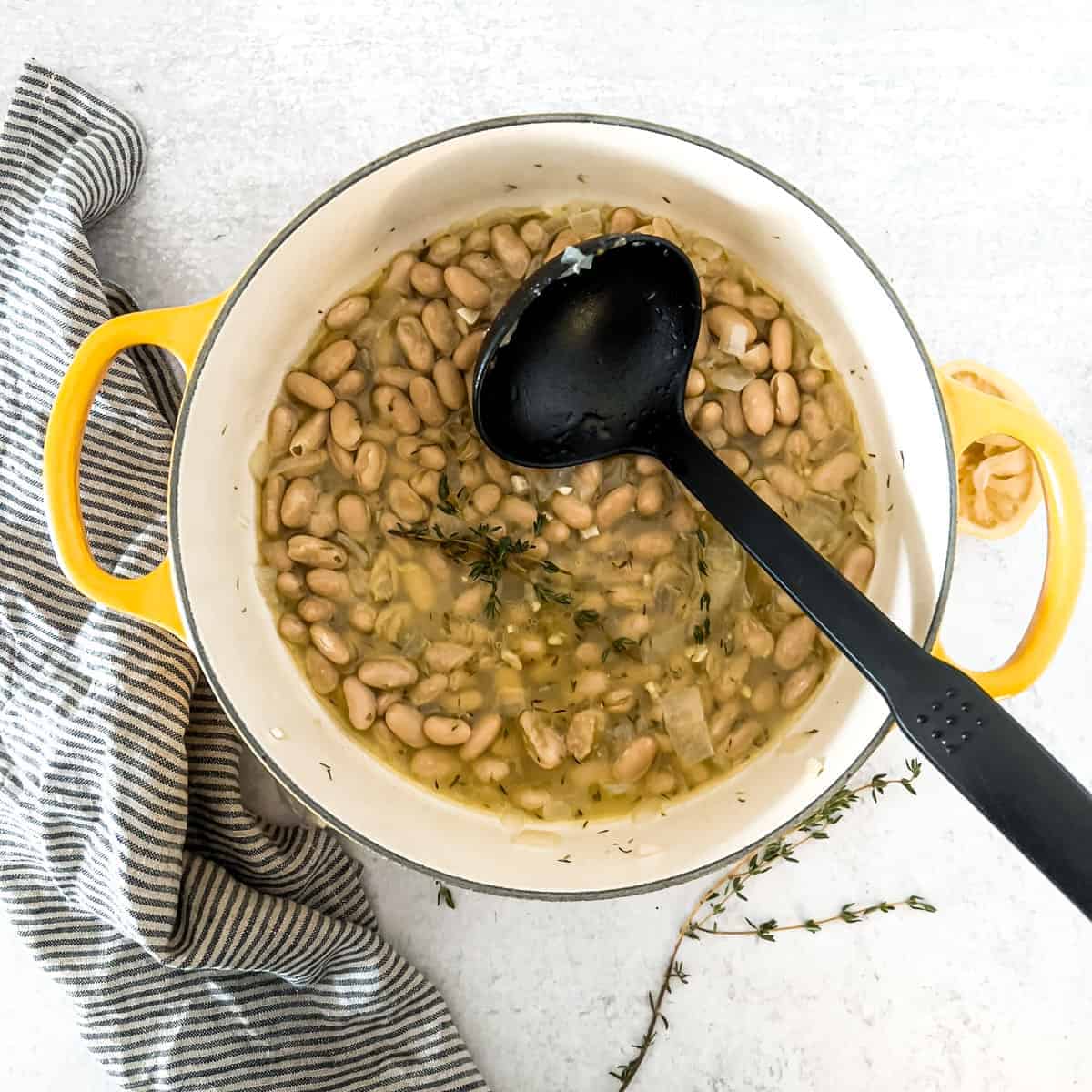 White bean stew cooking in yellow dutch oven and black ladle.