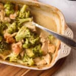 Baked chicken divan with serving spoon in small baking dish on wooden board.
