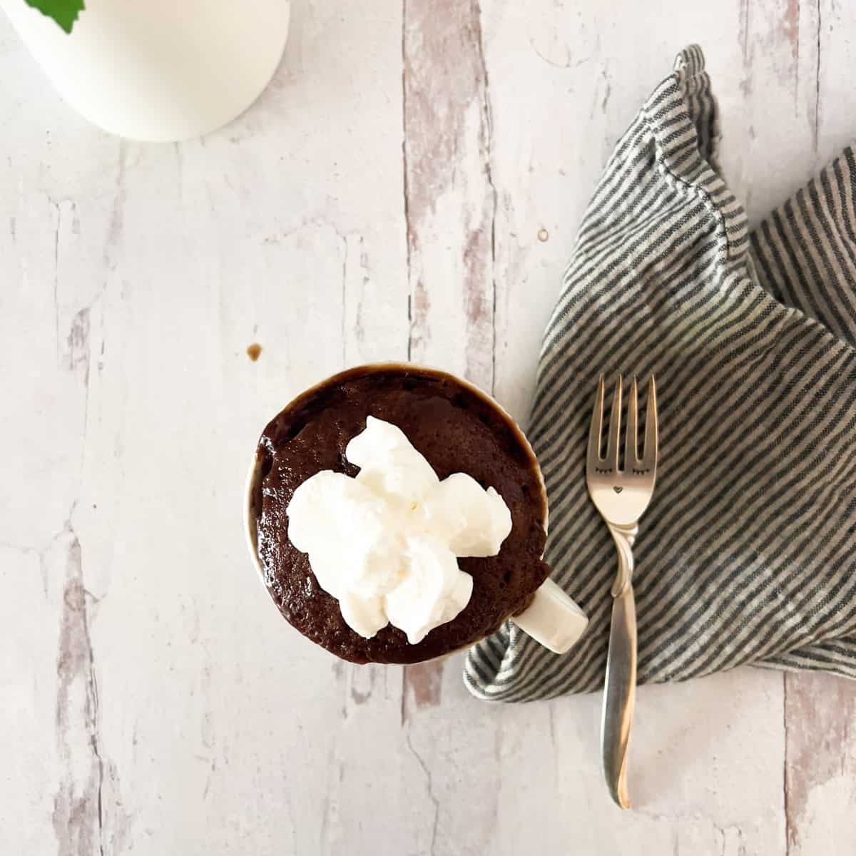 White mug filled with Nutella mug cake and whipped cream with striped napkin and fork on side.