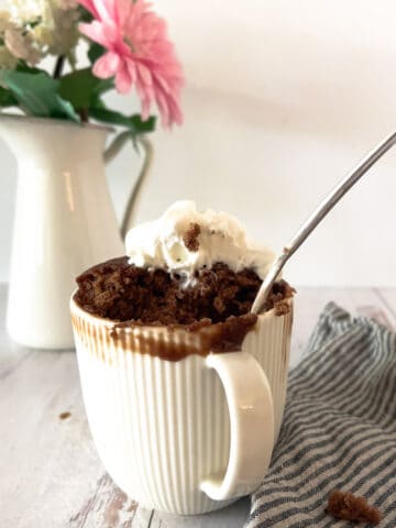 White mug filled with Nutella mug cake and whipped cream with striped napkin and fork on side and flowers in background.