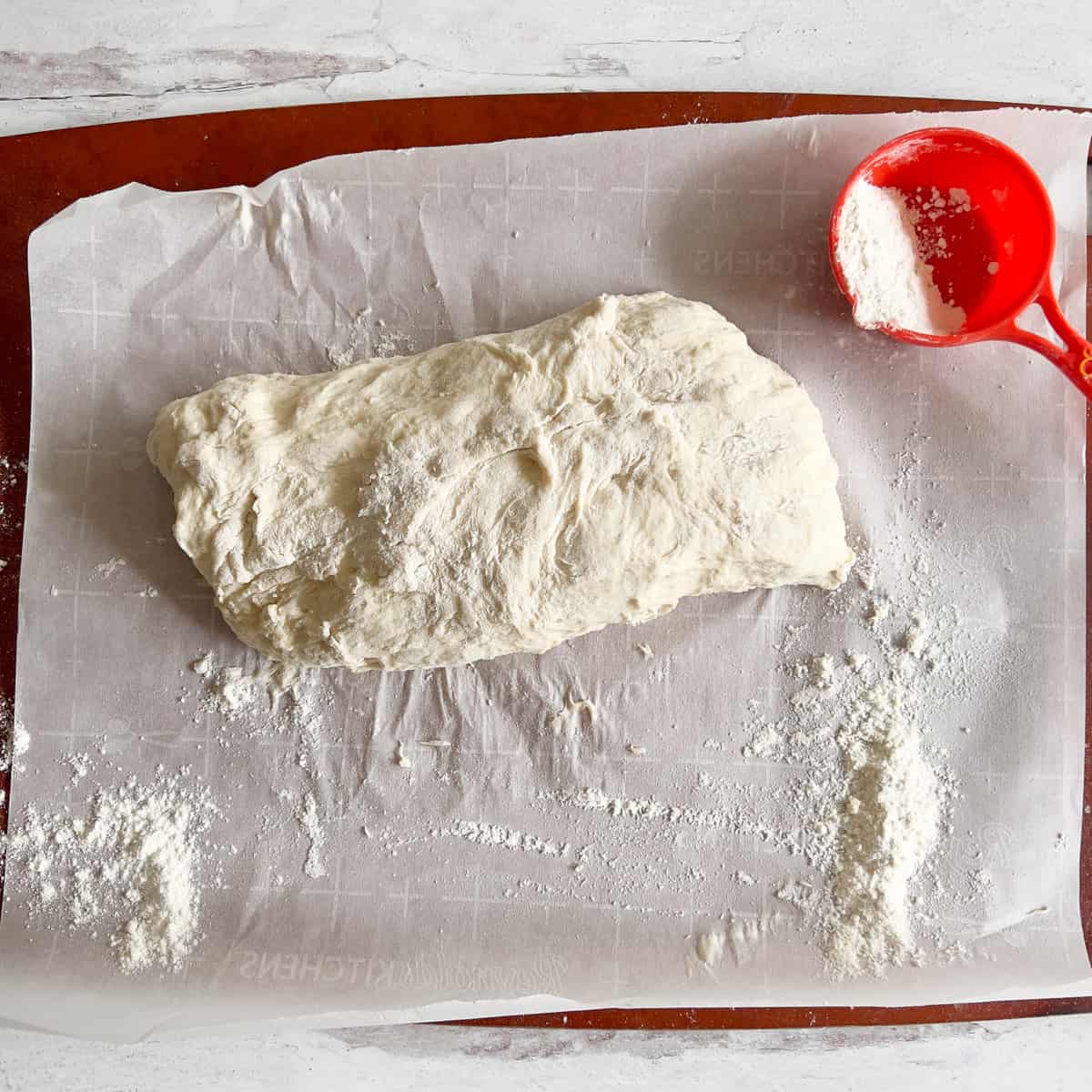 Ciabatta dough stretched out in a rectangle on parchment lines cutting board with orange measuring cup partially filled with flour.