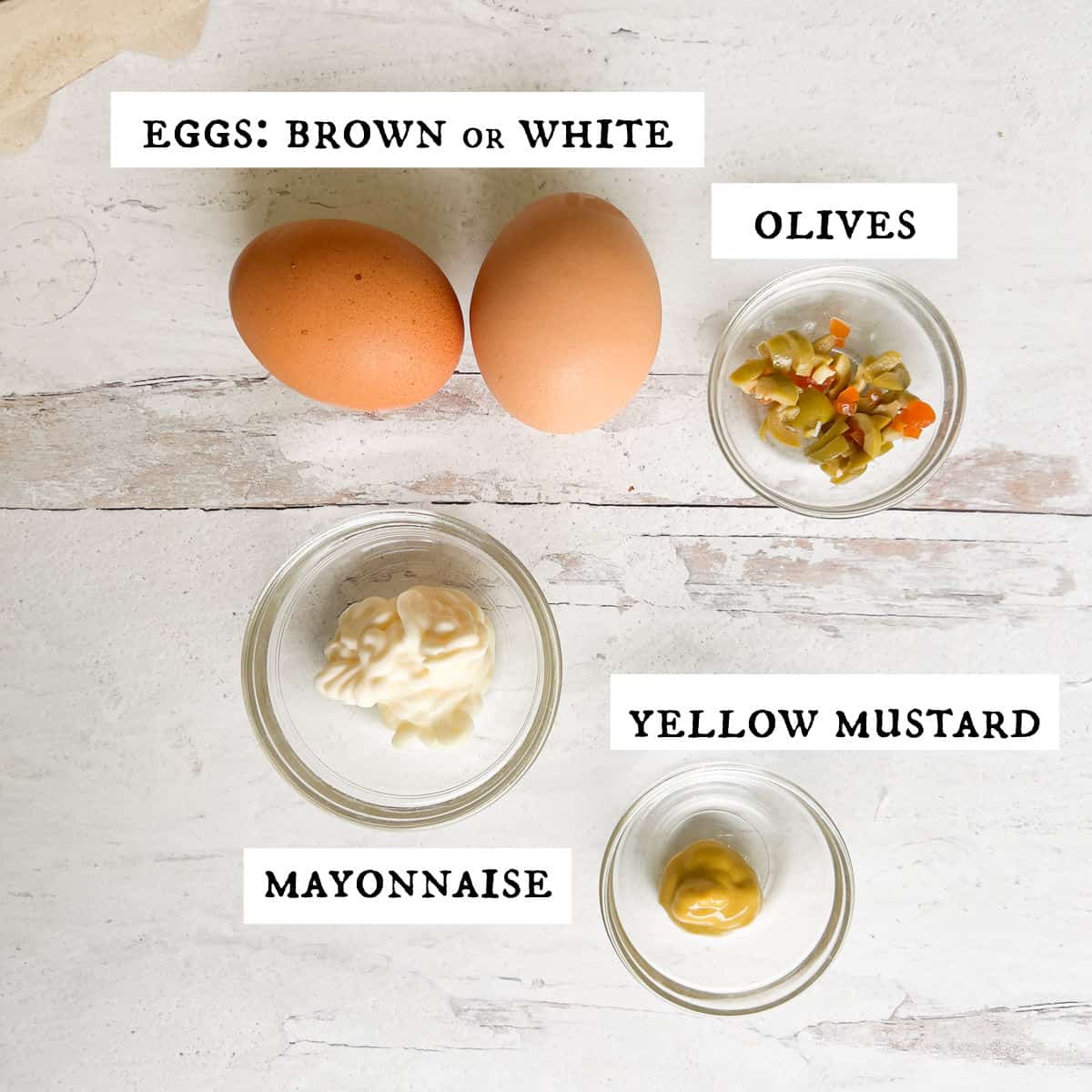 Two brown eggs, chopped olives, yellow mustard and mayo in bowls on white table.