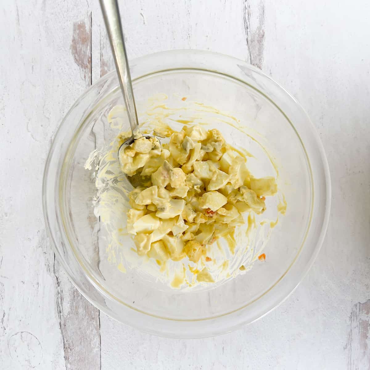 Chopped egg salad and spoon in clear bowl on white wood table.