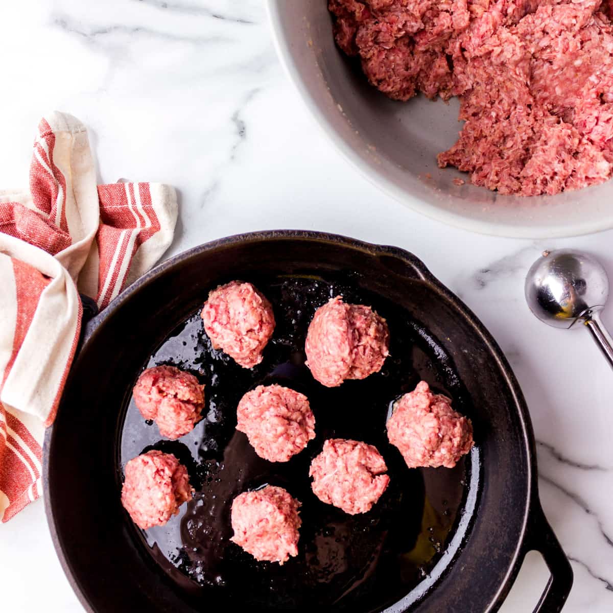 Meatballs frying in skillet with bowl of ground beef and cookie scoop on the side.