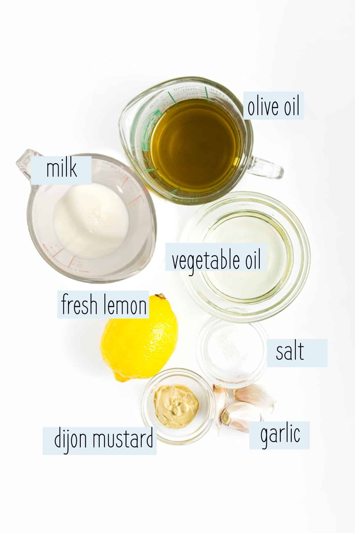 Milk, olive oil, garlic cloves, Dijon mustard and fresh lemon measured out of white surface.