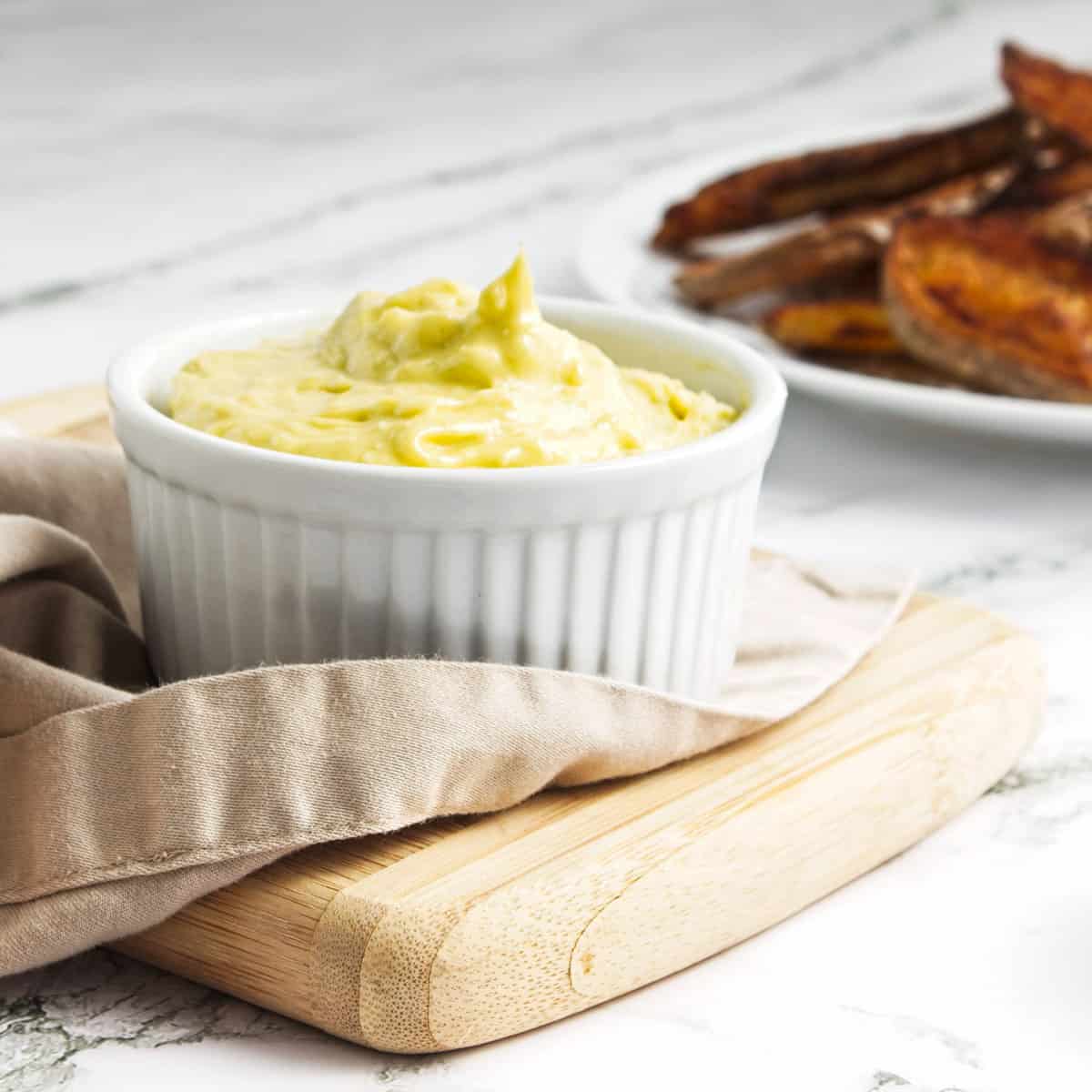 Garlic mayonnaise in small white ramekin on cutting board with plate of toasted bread in background.