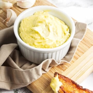 Garlic mayonnaise in small white ramekin on cutting board with toasted bread spread partially spread with the mayo.