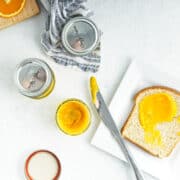 Small mason jars of orange curd next to a plate with slice of bread spread with the orange curd.