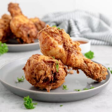 Two golden fried chicken drumsticks on grey plate with plate of more chicken in background.