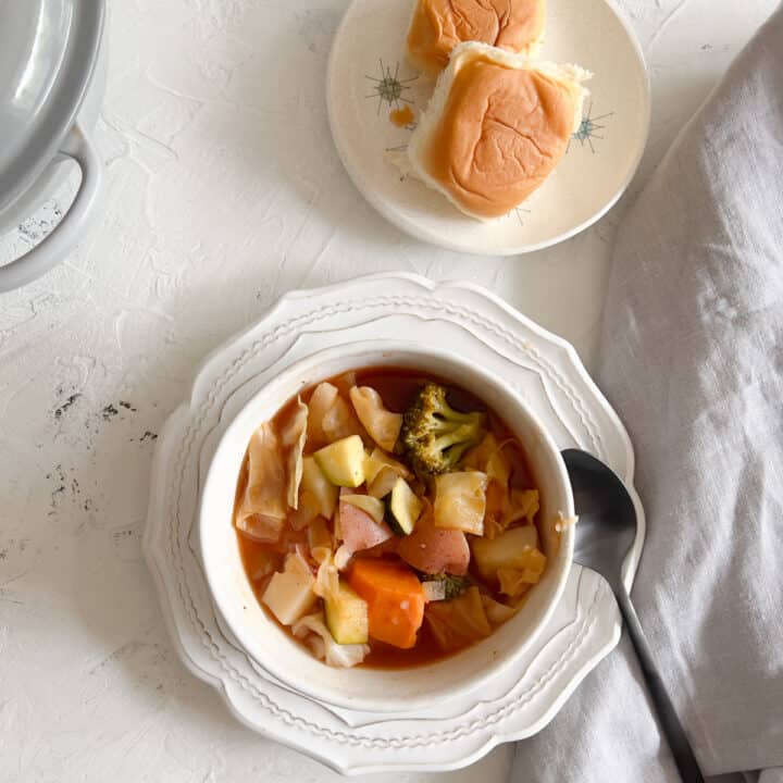 Bowl of V8 vegetable soup with soup spoon and plate of dinner rolls.