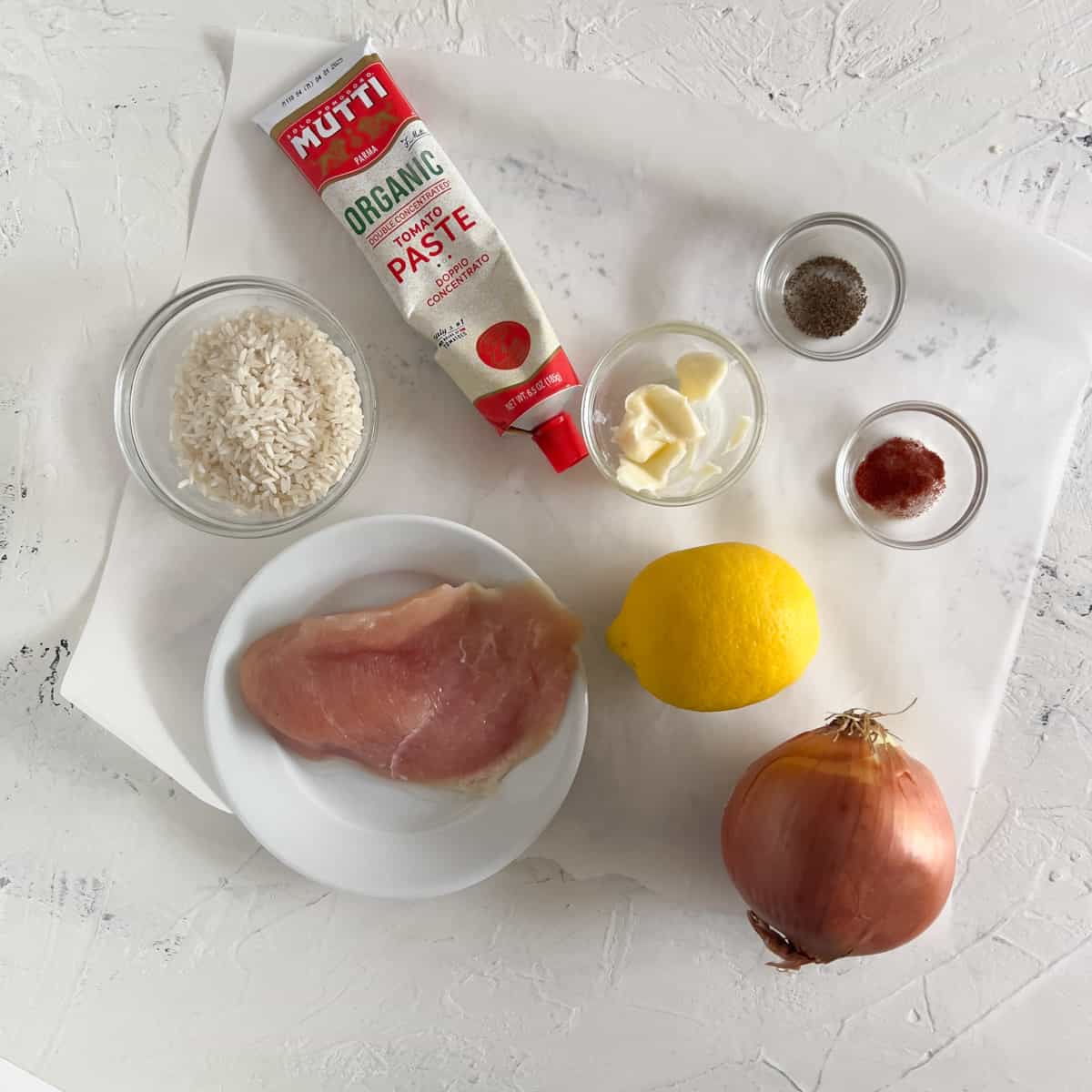 Lemon chicken ingredients: uncooked chicken breast rice, lemon, onion and tube of Mutti tomato paste on parchment paper.