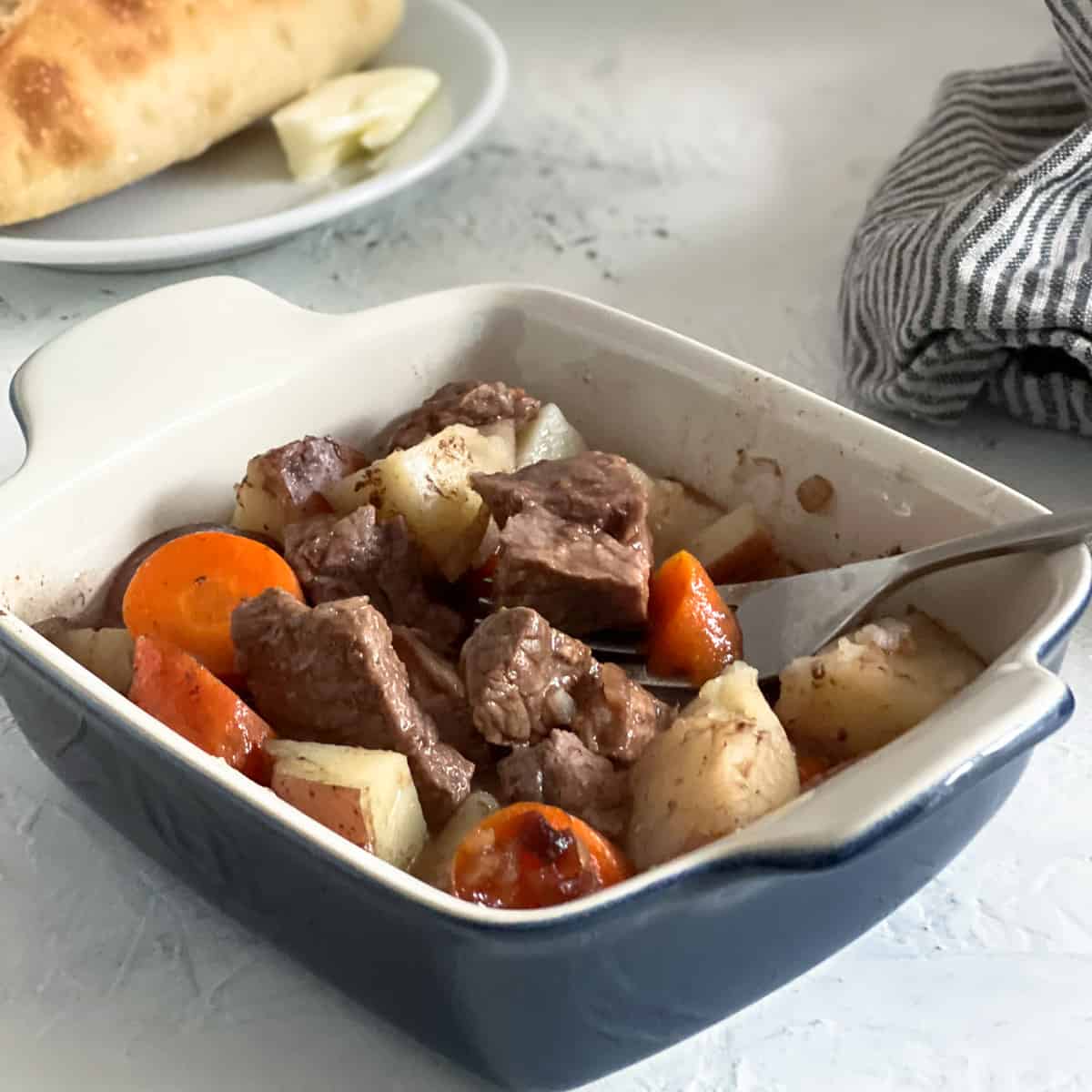 Baked beef stew casserole in blue baking dish with striped napkin and bread plate on side.