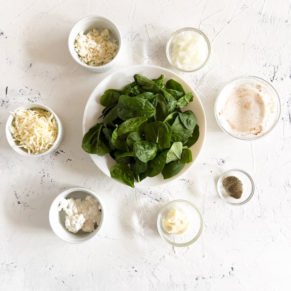 Bowl of baby spinach, and small bowls of feta cheese, cottage cheese, shredded mozzarella, heavy cream,. black pepper and diced onions.