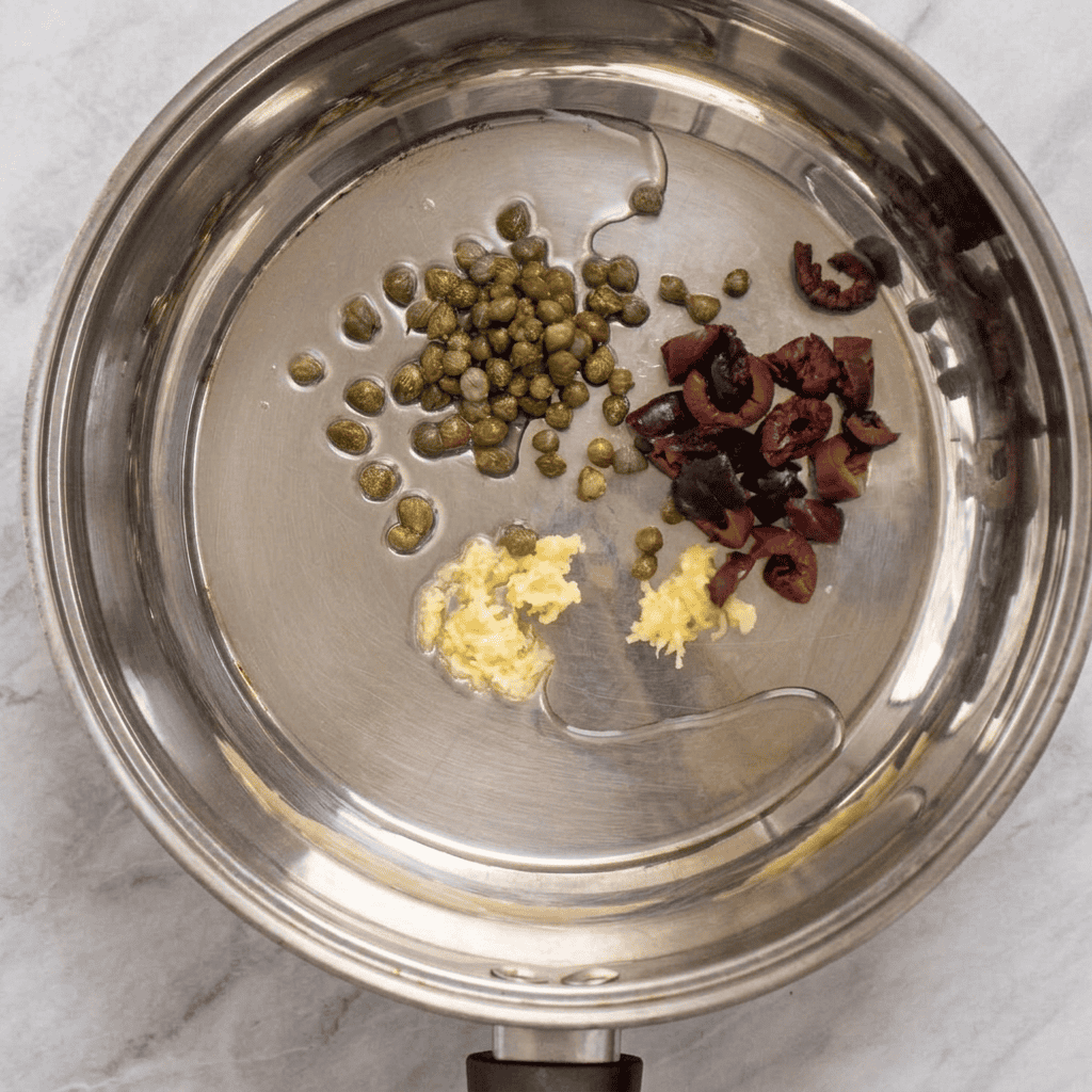 A stainless steel pan with chopped garlic, sliced black olives, capers, and a drizzle of olive oil on a marble surface—perfect ingredients for starting a flavorful pasta with olives and capers.