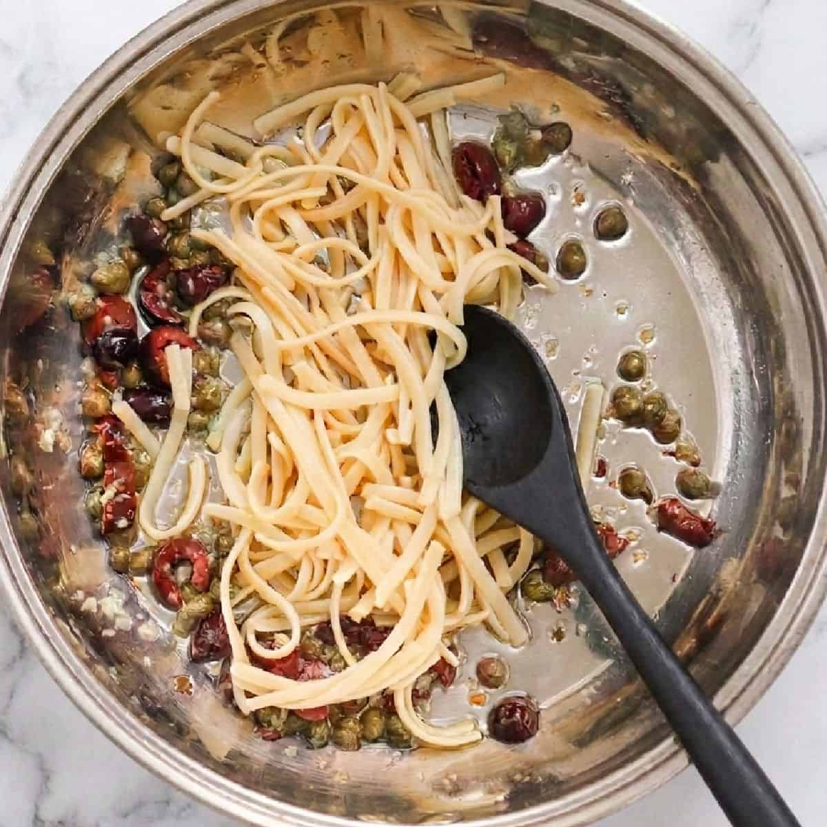 Cooked linguine, capers, and black olives come together in this delicious pasta with olives and capers, served in a stainless steel pan with a black spoon, on a marble surface.