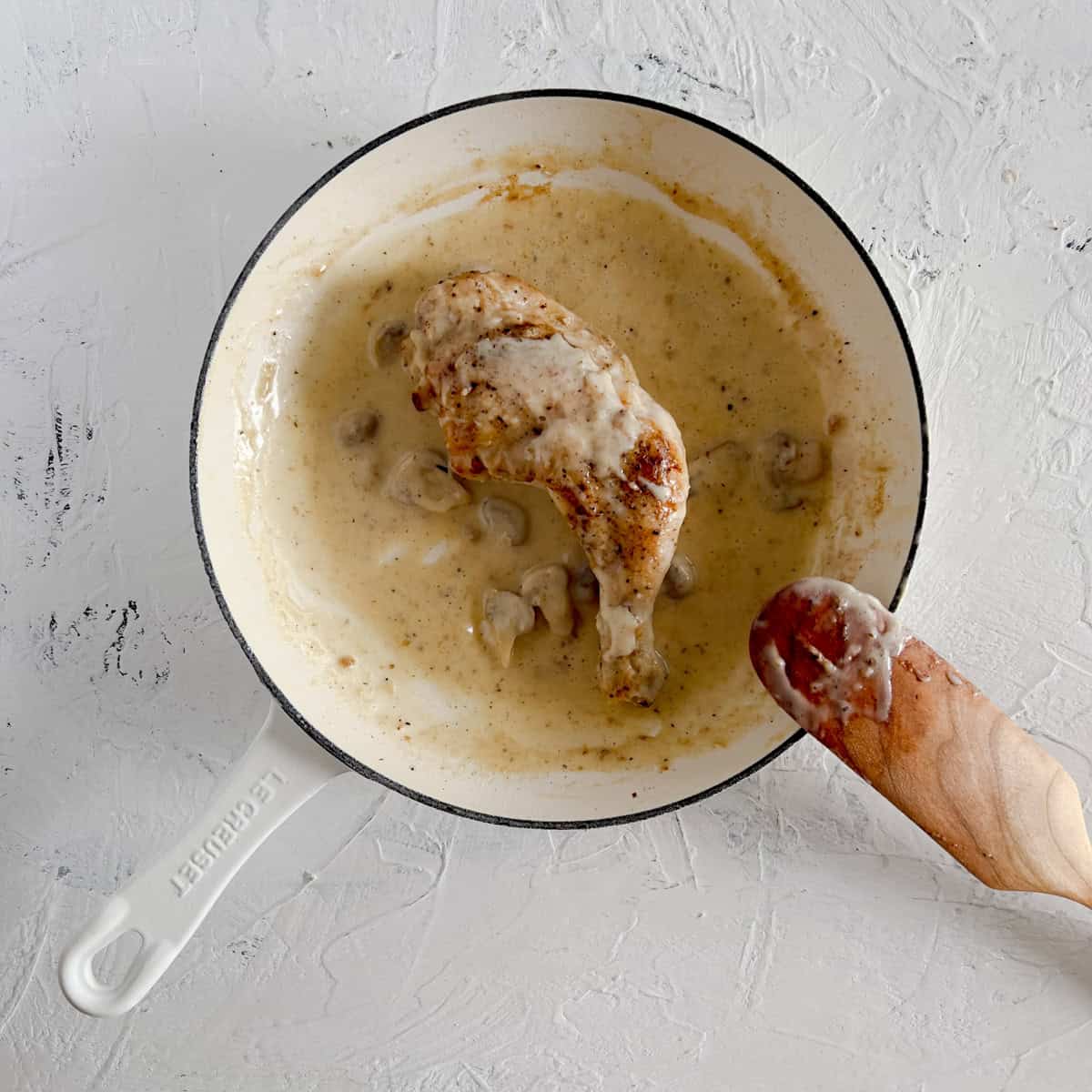 Chicken leg simmering in a mushroom sauce in a white skillet.