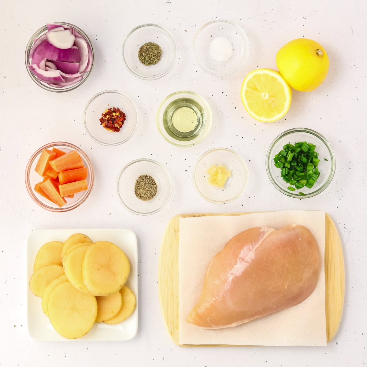 Raw chicken, carrots, onions, potatoes, garlic, lemon, and spices in clear bowls on table.