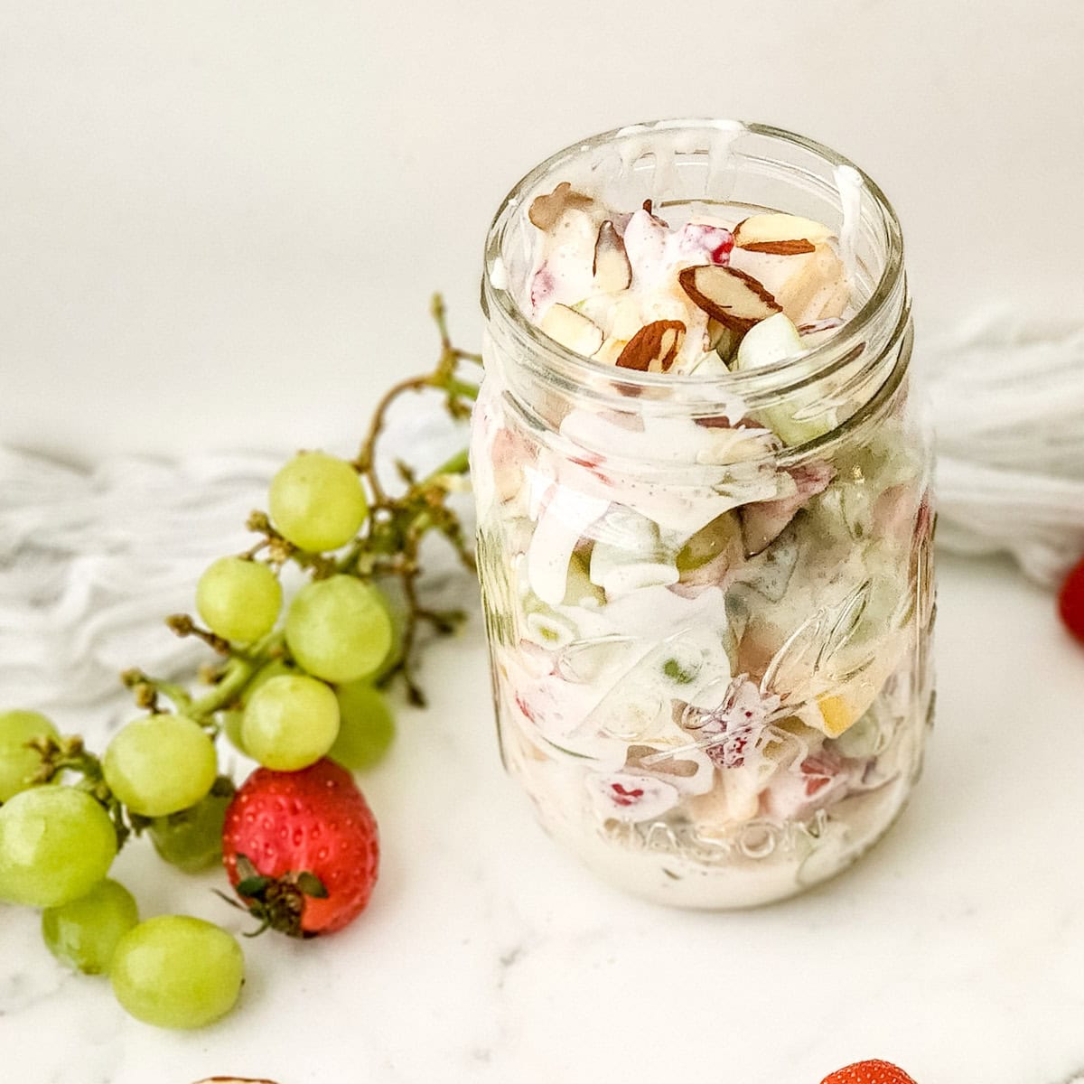 Creamy fruit salad in mason jar with fresh grapes and strawberries scattered on table.