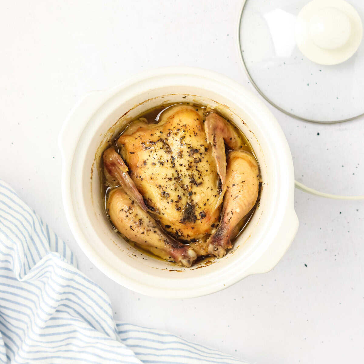 Whole cooked chicken seasoned with pepper in a white slow cooker, perfect for those Cooking for one or seeking clever tips and hacks, sits beside a striped cloth and glass lid.