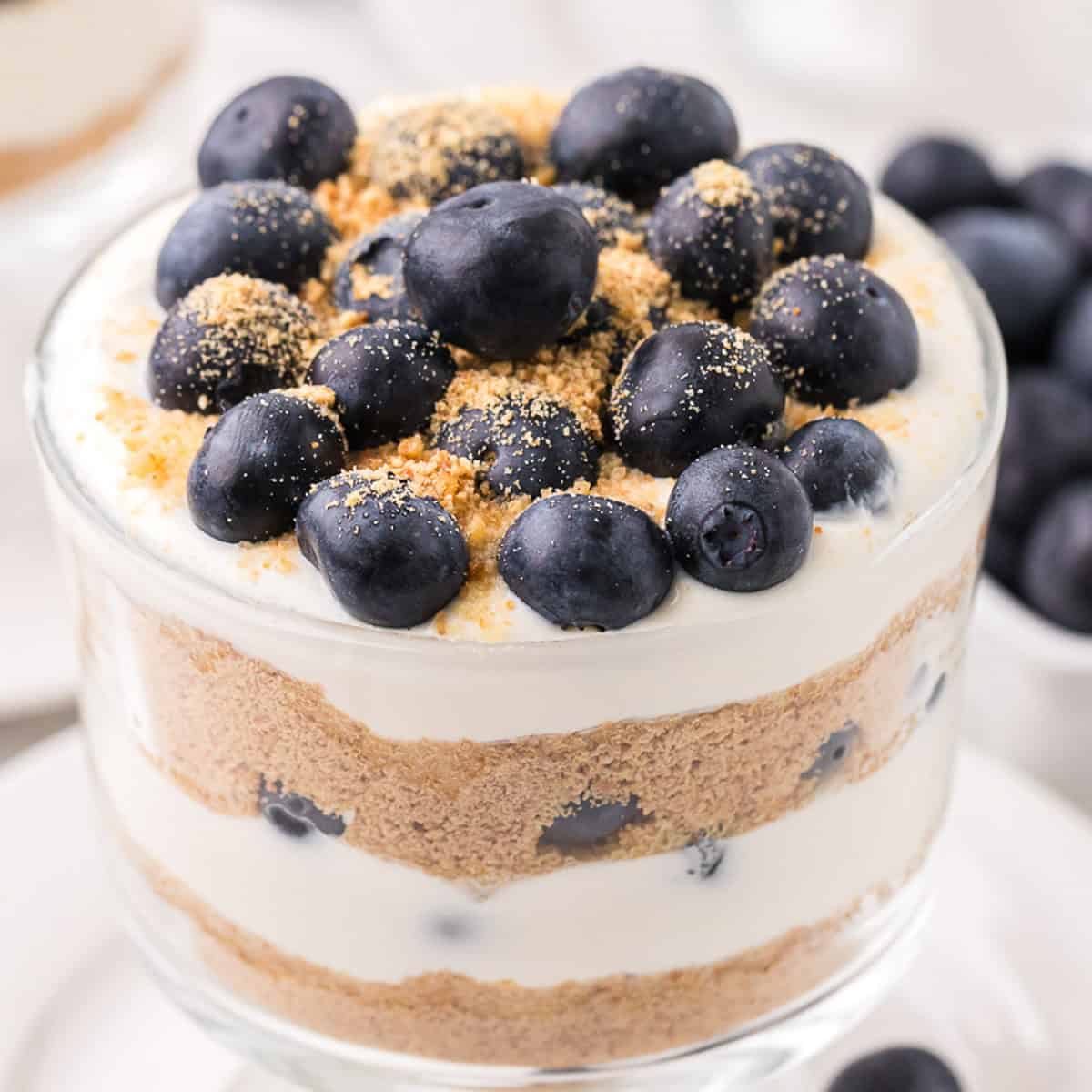 A layered dessert in a glass with blueberries, cream, and crumbled biscuit topping.