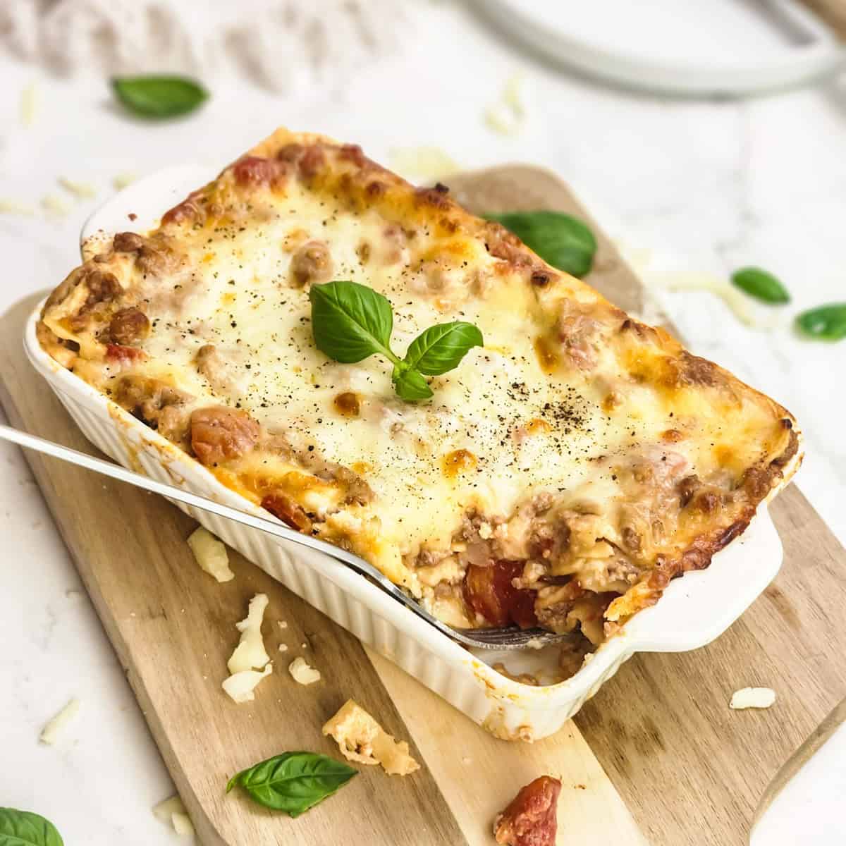 A baked mini lasagna in a white dish, topped with melted cheese and basil, on a wooden board.